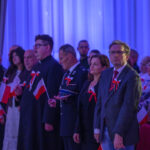 DSC_8040 Grupa ludzi w sali z niebieskim oświetleniem, trzymających polskie flagi. Mają przypięte czerwono-białe kokardy. Tło z zasłonami.