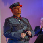 DSC_8047 Na zdjęciu mężczyzna w historycznym wojskowym mundurze, z mikrofonem w dłoni. Tło jest rozmyte, oświetlone na kolorowo. Obok widoczny jest drugi mężczyzna grający na trąbce.