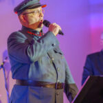 DSC_8048 Mężczyzna w granatowym mundurze z czapką śpiewa do mikrofonu. Tło w odcieniach fioletu. Obok widoczny statyw.