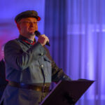 DSC_8052 Mężczyzna w mundurze wojskowym mówi do mikrofonu. Tło oświetlone jest na niebiesko i fioletowo, z lekkimi pionowymi pasmami. Na pierwszym planie stojak na nuty.