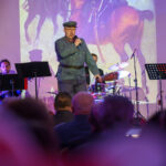 DSC_8059 Na scenie występuje osoba w mundurze, gra muzyka. Tło to obraz konia, a kolorystyka dominuje w ciemnych barwach. Widać kilka instrumentów muzycznych i publiczność.