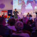 DSC_8063 Na scenie odbywa się koncert; artysta śpiewa, a zespół gra. W tle biało-czerwona flaga i obraz z żołnierzem i koniem. Publiczność siedzi na widowni.