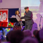 DSC_8066 Dwoje ludzi na scenie, mężczyzna w mundurze śpiewa do mikrofonu. W tle biało-czerwona flaga i herb Polski. Publiczność w cieniu, kolorowe oświetlenie.
