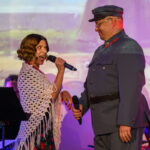 DSC_8070 Na scenie kobieta z mikrofonem i mężczyzna w mundurze, przy kolorowym tle. Kobieta nosi białą chustę. Mężczyzna ma szary mundur i czapkę.
