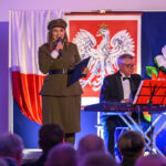 DSC_8078 Na scenie kobieta w mundurze mówi do mikrofonu, obok niej mężczyzna gra na keyboardzie. W tle znajduje się flaga Polski i godło. Dominuje czerwony, biały i niebieski kolor.