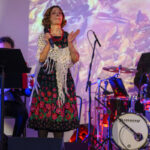 DSC_8082 Na scenie występuje kobieta w kolorowej sukience, obok perkusista. Tło jasne, abstrakcyjne wzory. Instrumenty i mikrofon na przodzie.