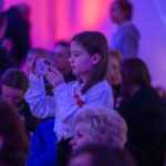 DSC_8085 Dziewczynka w białej bluzce fotografuje telefonem. Jasne tło w odcieniach różu i fioletu. W tle niewyraźne sylwetki ludzi.