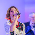 DSC_8089 Na zdjęciu młoda kobieta śpiewa do mikrofonu, ma na sobie kolorową sukienkę i biały szal. Tło jest fioletowo-niebieskie z oświetleniem scenicznym. Obok niej w oddali widać mężczyznę grającego na trąbce.