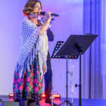 DSC_8092 Kobieta śpiewa na scenie, ubrana w folkową sukienkę z kwiatami i białą chustę. Tło jest niebieskie, z muzykami w tle. Światła sceniczne emitują ciepłe barwy.