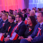 DSC_8094 Grupa ludzi siedzi w rzędu podczas wydarzenia, trzymając polskie flagi. Dominują kolory biały, czerwony i niebieski. Tło jest rozmyte ze względu na oświetlenie.