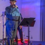 DSC_8098 Mężczyzna w niebieskim mundurze śpiewa na scenie. Tło jest oświetlone na niebiesko i czerwono. Obok mikrofon, w tle instrumenty.