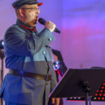 DSC_8099 Mężczyzna w mundurze wojskowym śpiewa do mikrofonu. Tło jest niebiesko-czerwone, obok stojak na nuty.