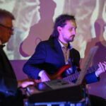 DSC_8101 Na zdjęciu trzej mężczyźni grają w ciemnych garniturach na instrumentach w jasnym pokoju. Ściana w tle ma kolorowe światła. Klawisze, gitara i sprzęt muzyczny są widoczne.