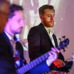 DSC_8104 Na zdjęciu widać muzyków na scenie w eleganckich garniturach. Mężczyzna z brodą gra na perkusji, drugi na gitarze. Tło jest jasne, z fioletowym oświetleniem.