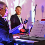 DSC_8108 Trzej mężczyźni w garniturach grają na keyboardzie i basie. Scena podświetlona jest na fioletowo. W tle stoją instrumenty perkusyjne.