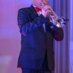 DSC_8113 Mężczyzna w ciemnym garniturze gra na trąbce. Tło jest rozmyte, w odcieniach niebieskim i różowym. Jasne oświetlenie podkreśla scenę.