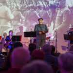 DSC_8117 Na zdjęciu widać scenę z grupą muzyków, w tym jedną osobę w wojskowym mundurze. Tło jest kolorowe, z historyczną projekcją. Widownia składa się z kilku osób, niektóre oświetlone punktowym światłem.