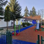 Plac zabaw z niebieską ścieżką i kolorową zjeżdżalnią. W tle białe budynki z czerwoną dachówką. Otoczone zielonymi drzewami.