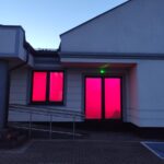 Budynek z szarymi ścianami, dwoma oknami z intensywnie różowym światłem. Na tle nieba o zmierzchu. Na dole widoczna rampa i bruk.