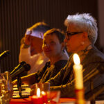 Trzy osoby, w tym jedna z jasnymi włosami, siedzą przy stole z mikrofonami. Na stole są świeczki, nadające ciepłe światło. Tło jest ciemne, co podkreśla atmosferę spotkania.