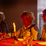 Trzy osoby siedzą przy stole w ciemnym pomieszczeniu. Stół pokryty jest czerwonym obrusem i oświetlony świecami. W tle ekran z rozmazanym obrazem.