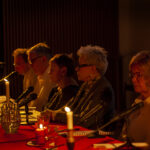 Zdjęcie pokazuje grupę osób przy stole z czerwonym obrusem, oświetlonych świecami. Stoły są ozdobione świecami w ciemnym pomieszczeniu. Osoby siedzą przy mikrofonach, co wskazuje na dyskusję lub spotkanie.