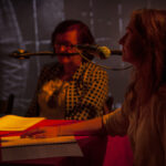 Dwóch ludzi siedzi przy stole z mikrofonem, w ciepłym świetle. Osoby skoncentrowane są na czytaniu dokumentów. W tle rozmyty obraz w odcieniach szarości.