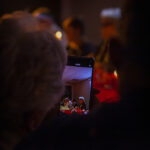 Na zdjęciu widać telefon komórkowy, na którym grupa osób siedzi przy stole. Tło jest ciemne, a scena oświetlona ciepłym światłem. Kolory dominujące to czerwień i czerń.