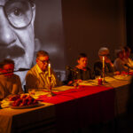 Na zdjęciu widać grupę osób siedzących przy stole, na którym palą się świece. Tło stanowi wielki czarno-biały portret. Atmosfera jest ciepła, z dominacją ciepłych odcieni.