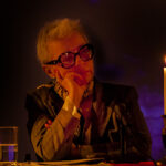 Osoba siedzi przy stole z dłońmi na twarzy. Ma szare włosy i okulary. Obok stoi zapalona świeca, a w tle ciemne oświetlenie.