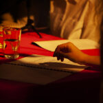 Ręka nad tekstem na czerwonym stole. Obok szklanki wody, delikatne światło, tło z zarysem osób. Scena o ciepłym tonie.