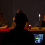 Na ciemnym tle widać osobę przy komputerze, przed nią palą się świece. W tle rozmyte postacie siedzą przy stole. Ciepłe, żółte światło dominuje na obrazie.