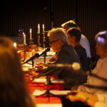 Grupa ludzi siedzi przy długim stole z czerwonym obrusem, oświetlonym świecami. Tło jest ciemne, a atmosfera intymna. Na stole widoczne są mikrofony i notatki.