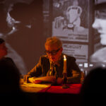 Na zdjęciu dwie osoby siedzą przy stole oświetlonym świecą. W tle wyświetlany jest czarno-biały film. Główne kolory to czerwony i brązowy.