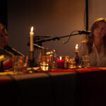 Na zdjęciu są trzy osoby przy stole z mikrofonami i świecami. Tło jest ciemne, światło pochodzi głównie od świec. Atmosfera jest intymna i ciepła.