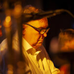 Nie rozpoznaję osób na zdjęciu. Obraz przedstawia mężczyznę w okularach, w białej koszuli, w ciepłym, żółtym świetle. W tle znajduje się niewyraźnie zarysowana postać, a atmosfera jest przytulna i intymna.