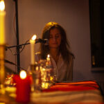 Osoba przy mikrofonie, w ciepłym oświetleniu świec. Tło ciemne, z delikatnym żółtym blaskiem. Przód stołu zdobią czerwone i białe świece.