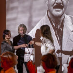 Na zdjęciu są trzy osoby w eleganckich strojach. Wręczają sobie białe torby pod czarno-białym portretem mężczyzny w tle. Dominuje czerwono-brązowe oświetlenie.