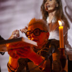 Na pierwszym planie osoba z białymi włosami i okularami trzyma książkę. Obok pali się świeca, a tło jest zaciemnione. Za nią widać kolejną postać.