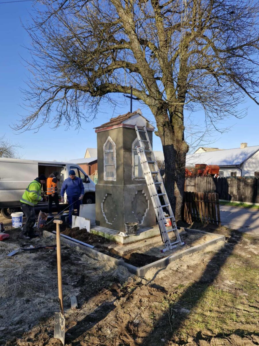 Na zdjęciu ludzie pracują przy małej kapliczce obok drzewa. Kapliczka jest szara, z drabiną opartą o nią. W tle jasnoniebieskie niebo i samochód dostawczy.
