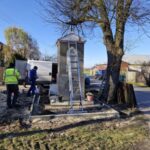 Na zdjęciu widać szary pomnik z drabiną na tle błękitnego nieba i dużego drzewa. Obok pracują dwaj mężczyźni w jaskrawych kurtkach. W tle stoi biały samochód dostawczy.