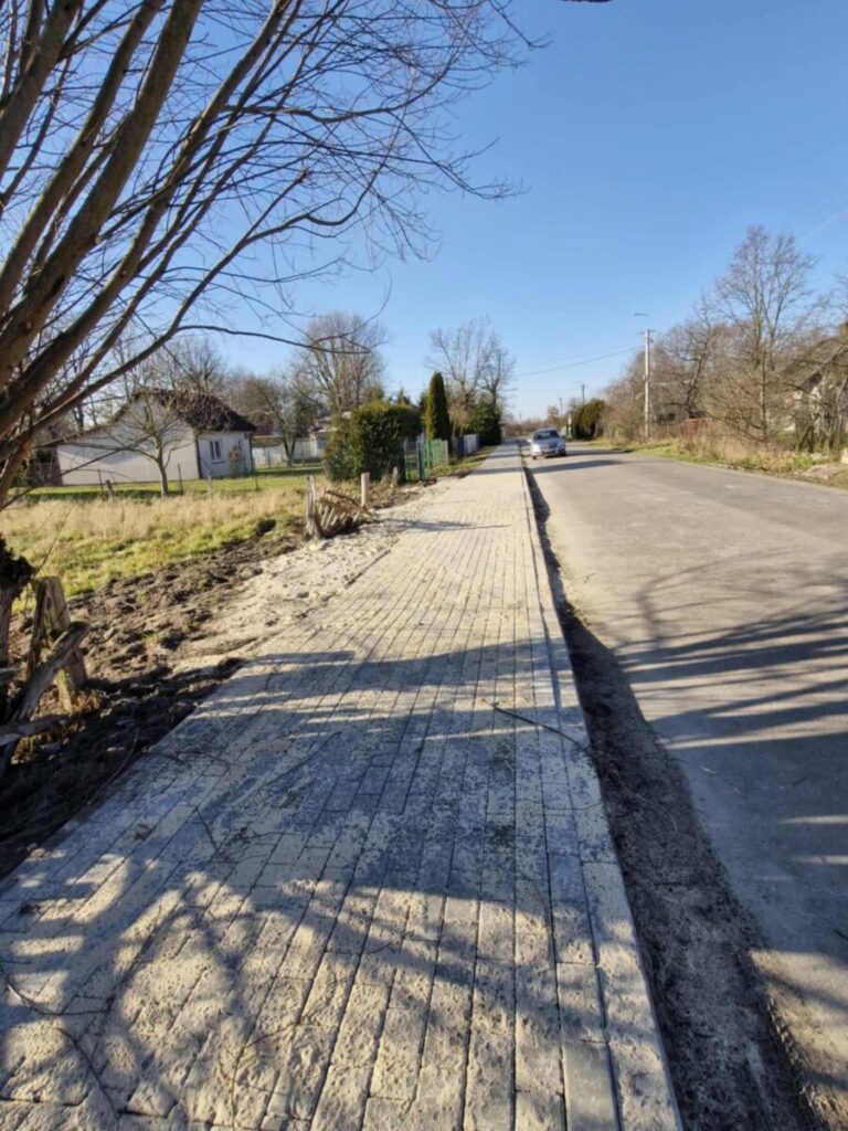 Chodnik z kostki brukowej biegnie wzdłuż drogi asfaltowej, po lewej stronie drzewa i trawnik. Słoneczny dzień, jasnoniebieskie niebo. W tle widoczny zaparkowany samochód i domy.