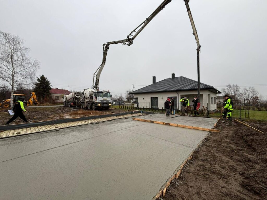 Na zdjęciu widać plac budowy z betonowaną powierzchnią. Ludzie w odblaskowych ubraniach, ciężarówka i dom w tle. Dzień jest pochmurny.