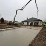 577061558_877181064657416_1952168253548782806_n Na zdjęciu widać plac budowy z betonowaną powierzchnią. Ludzie w odblaskowych ubraniach, ciężarówka i dom w tle. Dzień jest pochmurny.