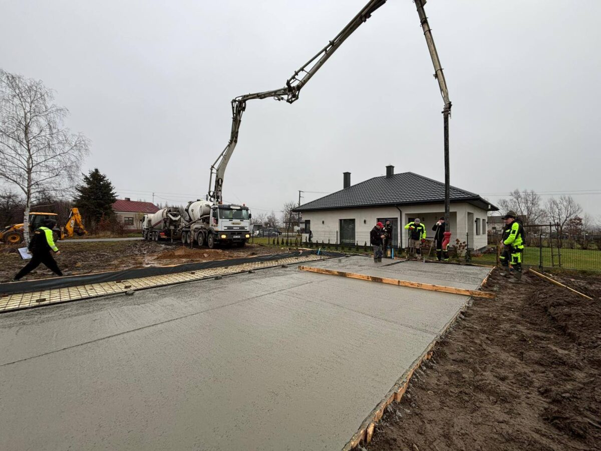 Na zdjęciu widać plac budowy z betonowaną powierzchnią. Ludzie w odblaskowych ubraniach, ciężarówka i dom w tle. Dzień jest pochmurny.