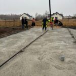 594177046_877180917990764_3816648569714731792_n Grupa pracowników buduje fundamenty z betonu. Noszą odzież ochronną, a w tle są domy i pola. Niebo jest szare i zachmurzone.