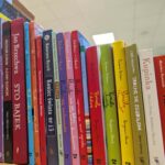 597199379_863142016110172_5126479399790451310_n Na obrazie widoczne są kolorowe grzbiety książek ustawionych na półce. Książki mają różnorodne kolory i są ustawione pod kątem. Tło jest rozmyte, z widocznym biało-kremowym sufitem.