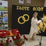 Scena na zdjęciu przedstawia uroczystość Złote gody. Z tyłu widać czarną tablicę z żółtymi obrączkami i napisami, otoczoną dużymi kwiatami. Na pierwszym planie kobieta w jasnej sukni przemawia do mikrofon