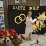 Kobieta w jasnej sukience przemawia przy mikrofonie na tle dekoracji „Złote Gody”. Po lewej stronie widać baner z krajobrazami gminy Dobre. Na stole stoją czerwone pudełka, otoczone bukietem czerwonych róż.