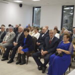 Grupa elegancko ubranych osób siedzi na krzesłach w jasnej sali. Ściany są białe, przez okna widać drzewa. Na stole z prawej strony są talerze i filiżanki.